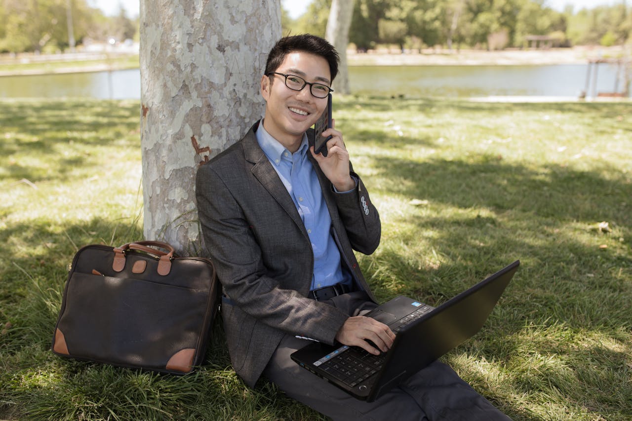 Asian man with glasses sitting under a tree working on a laptop